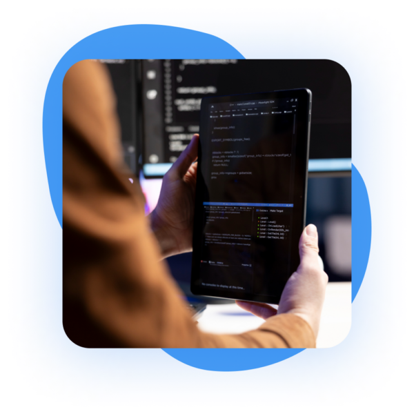 Person holding a tablet displaying lines of code, blurred computer screens in the background, all within a blue frame.
