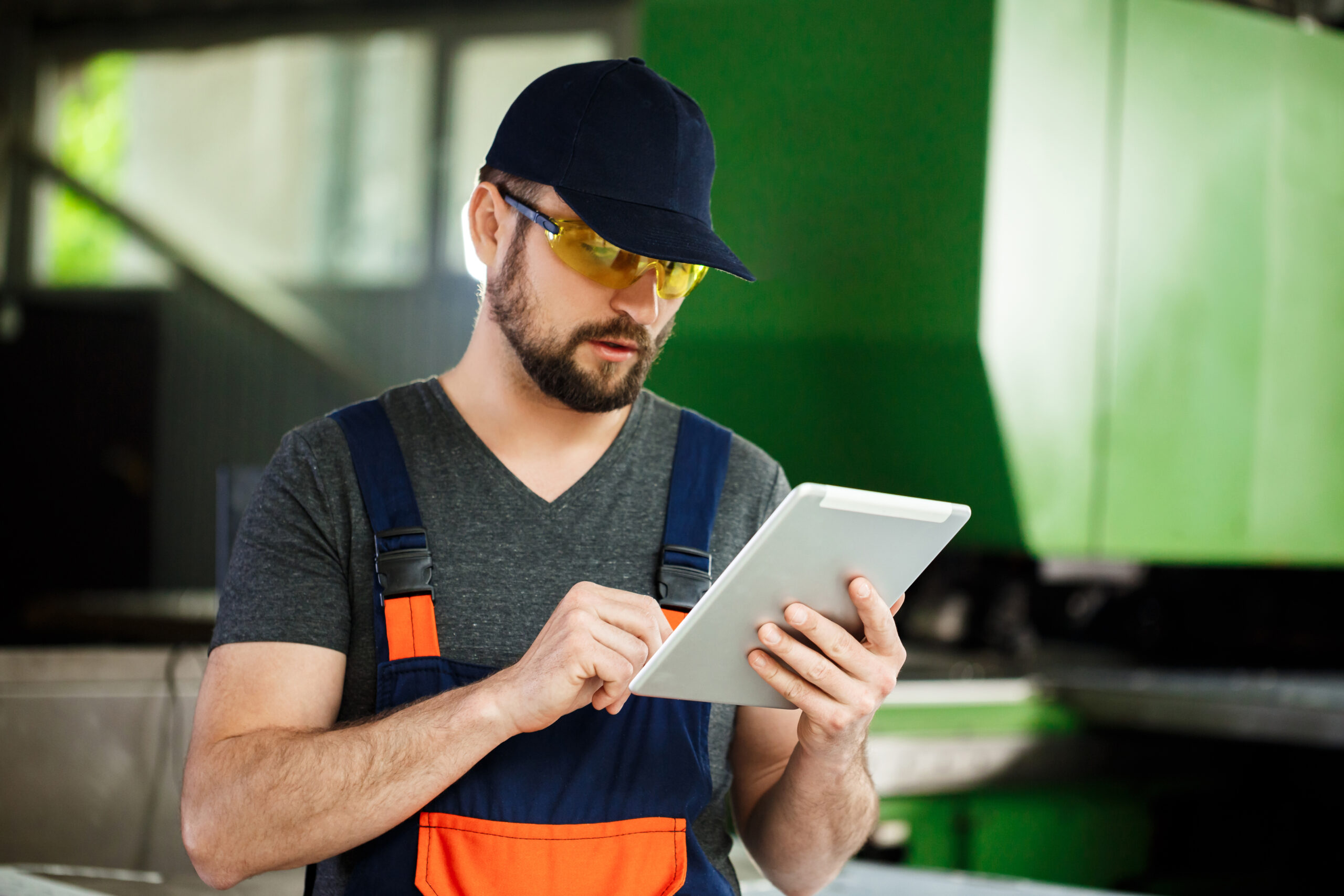 Focused male worker in a blue cap, safety glasses, and overalls using a tablet in an industrial setting.