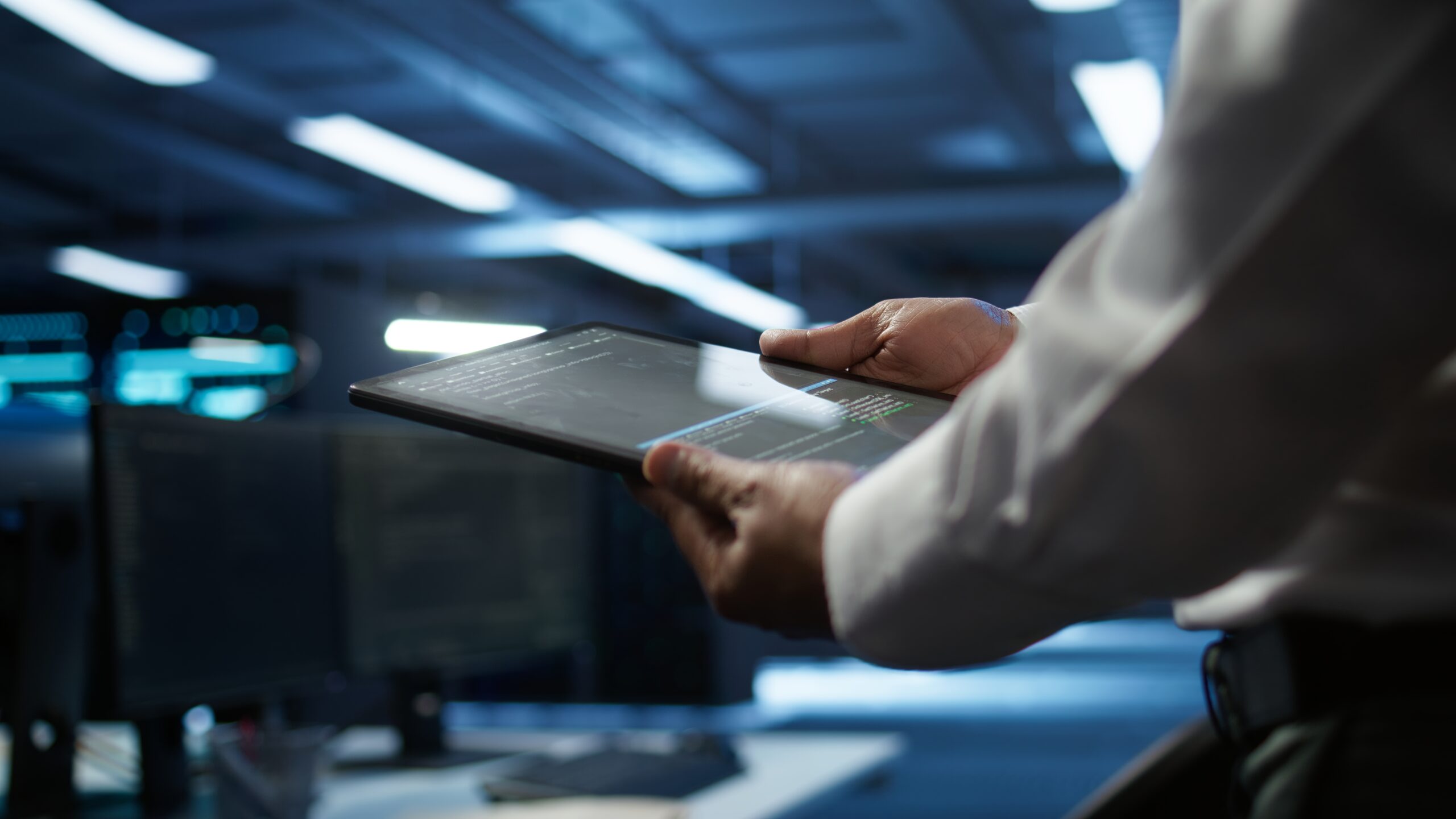 A person in a white shirt holding a tablet displaying code in a dimly lit, high-tech server room.