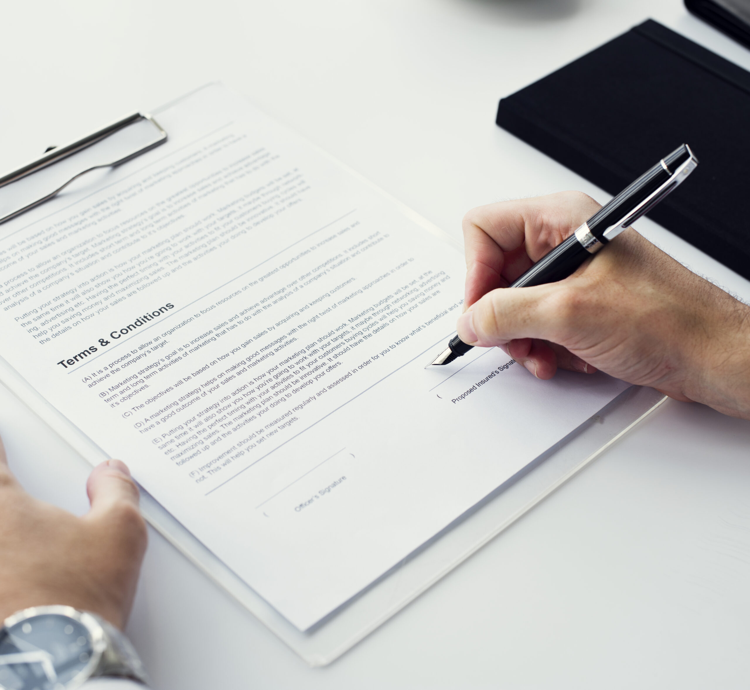 A person signing a document titled "Terms & Conditions" with a black pen on a white surface, with a watch and notebook visible.